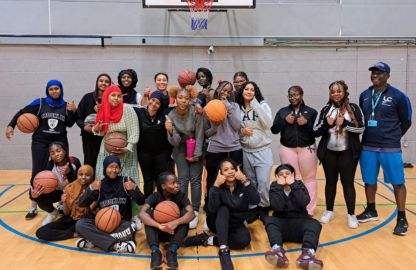 Group of diverse girls basketball players and coach at Lambeth College posing together in a gymnasium, with some holding basketballs and all smiling at the camera, standing and kneeling in two rows. A basketball hoop is visible in the background.